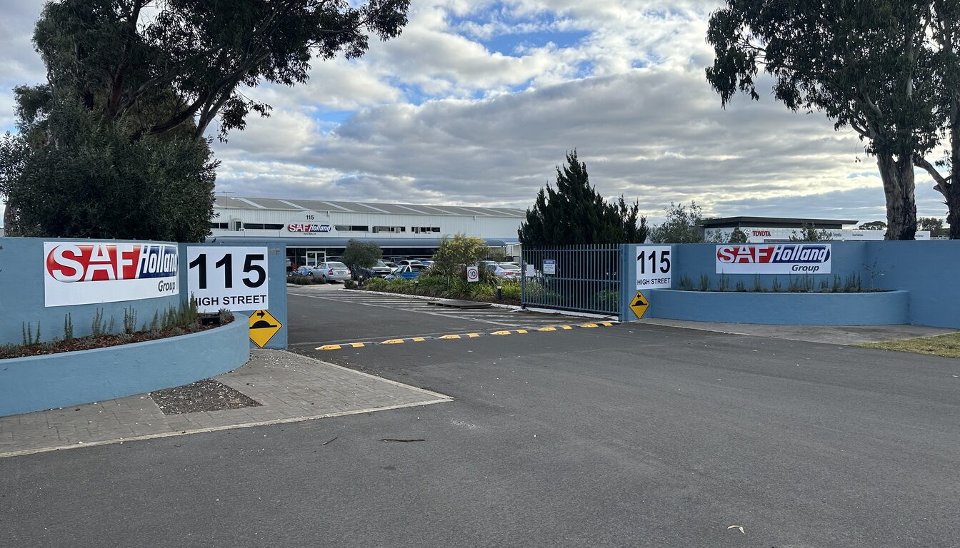 Industrial site entrance with two company signs and 115 High Street number signs on curved blue walls either side of a driveway, with a yellow speed hump warning sign and a speed limit sign