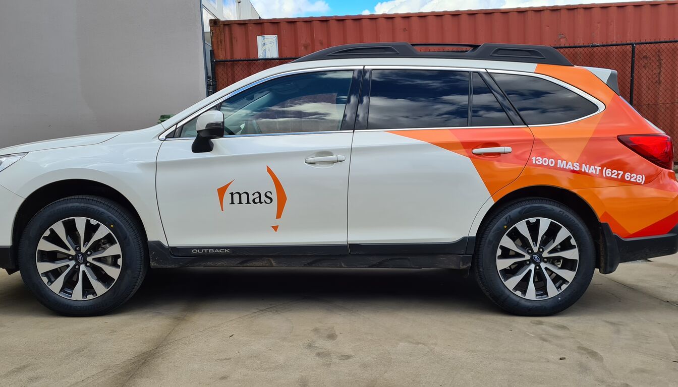 White station wagon parked in a yard with a partial vinyl wrap: a logo on the front door and orange geometric graphics with a phone number across the rear doors and quarter panel