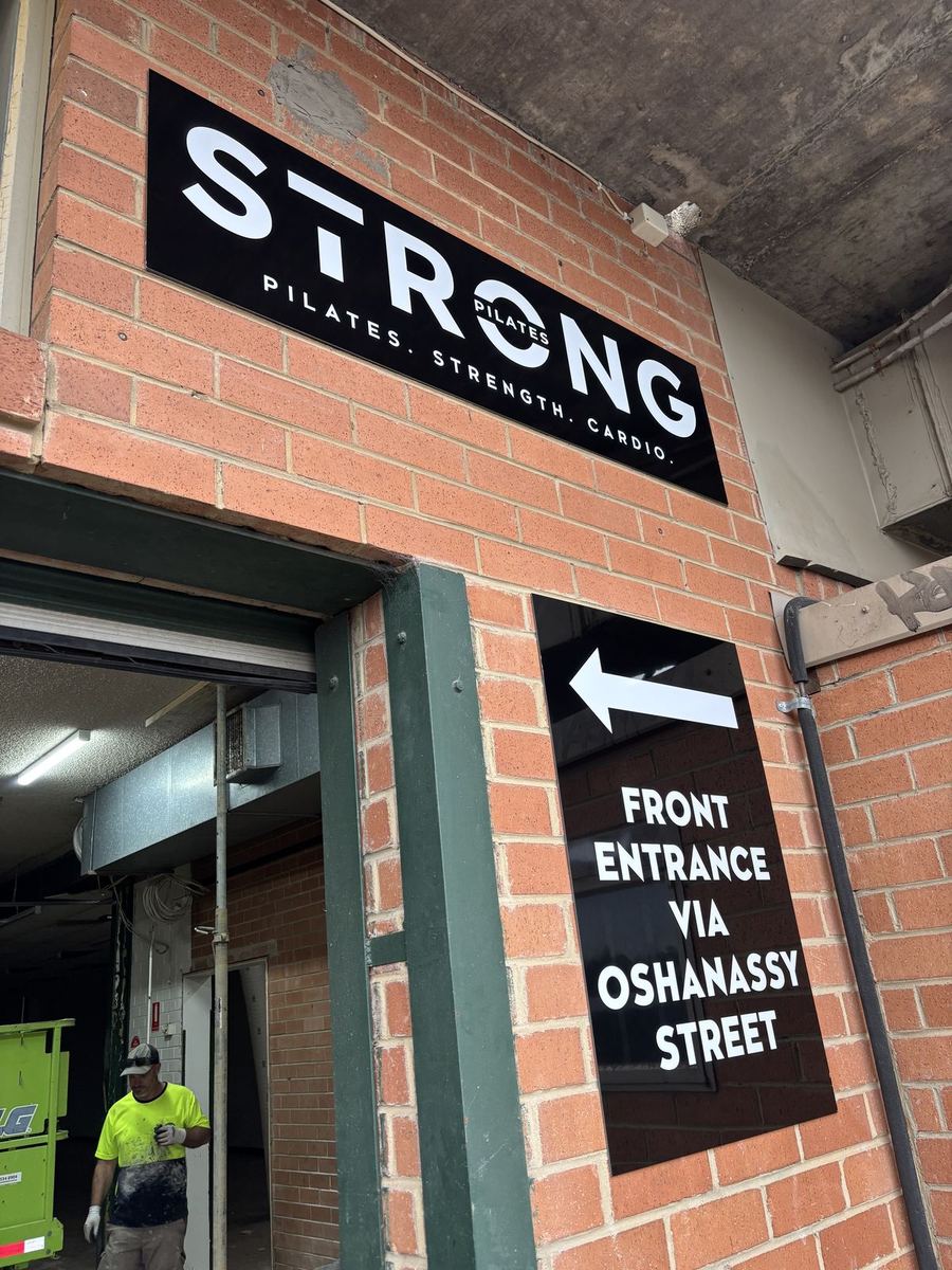 Black directional panel with a white arrow to the front entrance, on a brick wall below a name sign