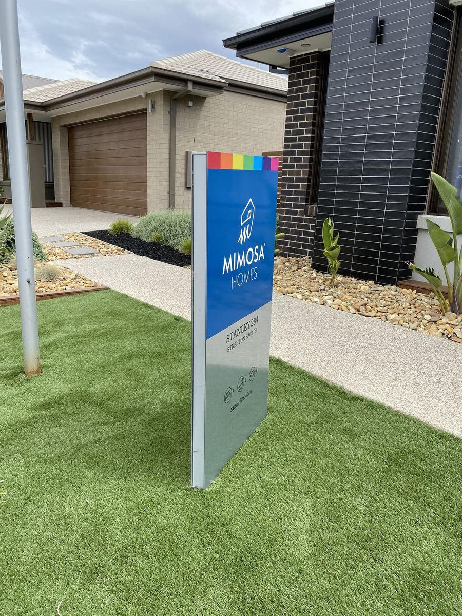 Freestanding blue and brushed metal pylon sign identifying a display home by name on its front lawn